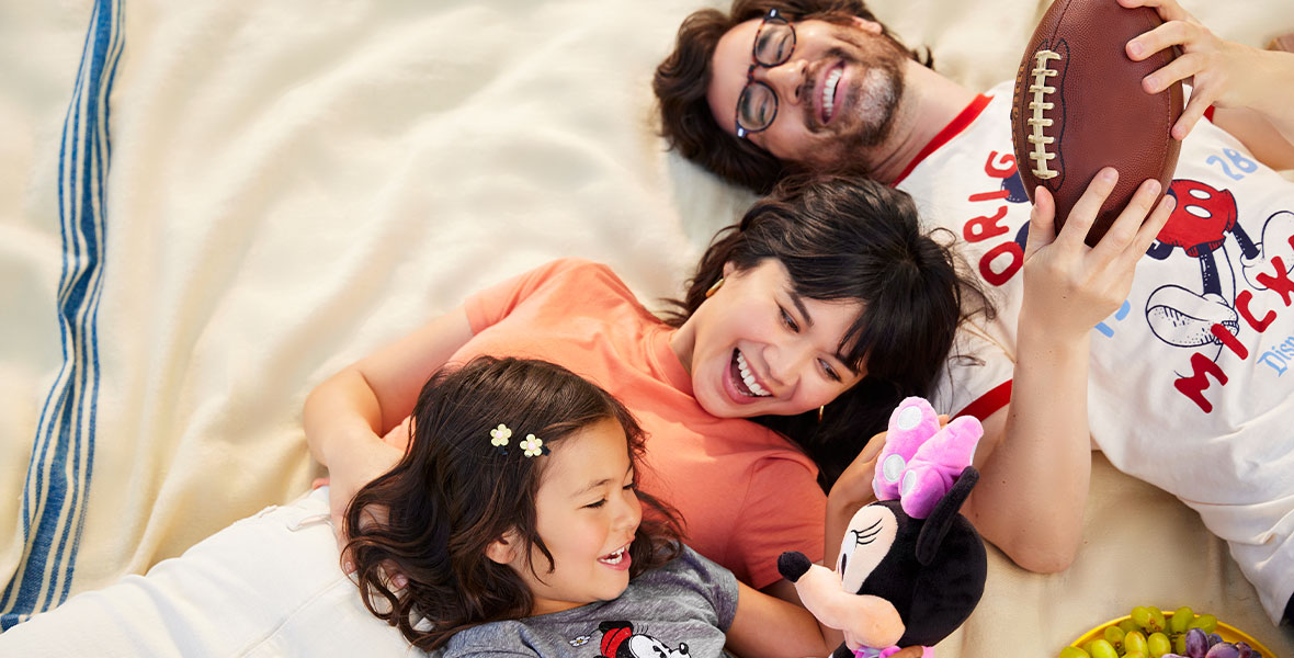 Two adults and a child lay on a blanket, laughing and smiling. The child is holding and looking at a Minnie Mouse plush, while the adult in the upper right corner holds a football.