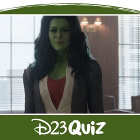 She-Hulk (Tatiana Maslany) stands in a courtroom. Her skin is bright green, and she has long wavy blue hair. She is wearing a red shirt with a white button up, and a gray blazer. The jury behind her is ducking behind the wood partition.