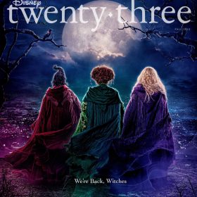 The front cover of Disney twenty-three, featuring the publication logo above the backs of Sarah, Winnifred, and Sarah Sanderson, all wearing cloaks and standing on a wooded hill overlooking a town at night. The text below them states, “We’re back, witches.”