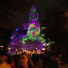 Carthay Circle lit up with neon green, pink and purple colors. Skeleton holding up marquee reading Mister Oogie Boogie with D23: The Official Disney Fan Club written on both sides of the skeleton in yellow. Trees surrounding Carthay Circle are lit up with purple lights.