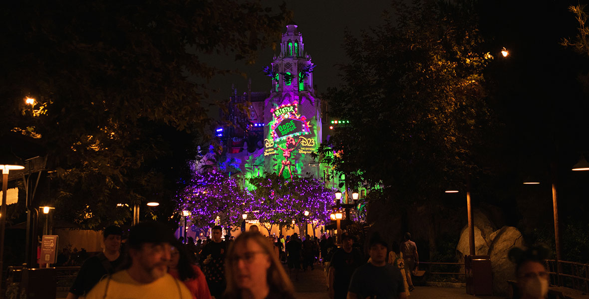 Carthay Circle lit up with neon green, pink and purple colors. Skeleton holding up marquee reading Mister Oogie Boogie with D23: The Official Disney Fan Club written on both sides of the skeleton in yellow. Trees surrounding Carthay Circle are lit up with purple lights.
