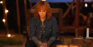 Reba McEntire, in character as Sunny Barnes, wears a denim shirt and sits in a wooden rocking chair in front of a bonfire. She has red, shoulder-length hair and a pensive look on her face.