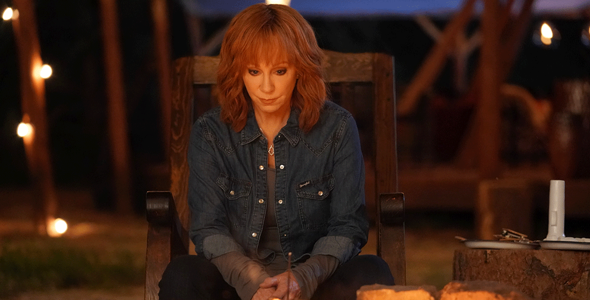 Reba McEntire, in character as Sunny Barnes, wears a denim shirt and sits in a wooden rocking chair in front of a bonfire. She has red, shoulder-length hair and a pensive look on her face.