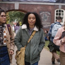 Belissa Escobedo in character as Izzy, Whitney Peak in character as Becca, and Lilia Buckingham in character as Cassie. The characters are dressed for fall, wearing sweaters and jackets, and they are walking out of school next to a crowded bike rack.