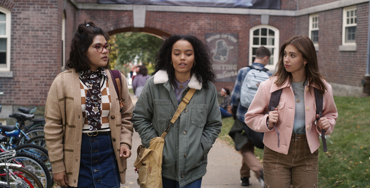 Belissa Escobedo in character as Izzy, Whitney Peak in character as Becca, and Lilia Buckingham in character as Cassie. The characters are dressed for fall, wearing sweaters and jackets, and they are walking out of school next to a crowded bike rack.