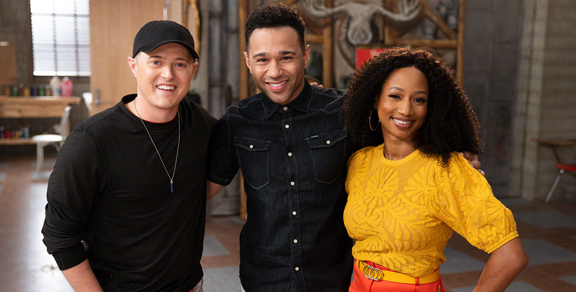 (From left to right): Lucas Grabeel, Corbin Bleu, and Monique Coleman pose for a group photo on the set of High School Musical: The Musical: The Series Season 4. Grabeel wears a black hat, a black shirt, a black watch, and gray jeans, and completes his look with a pendant necklace. Grabeel wears a denim blue button-up shirt and white jeans. Coleman wears a short-sleeved, yellow floral top, a yellow belt, and an orange skirt. All three are smiling.