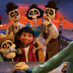 Miguel from Coco stands wearing a red hoodie over a white tank top. His ancestors portrayed by skeletal figures, are standing around him in traditional Mexican clothing.