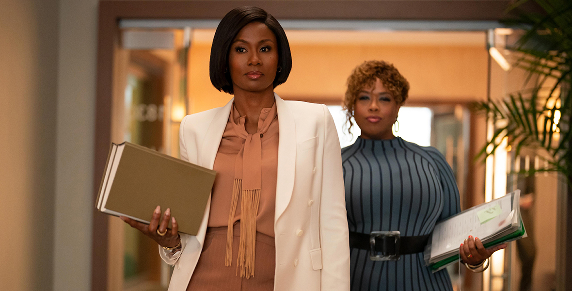 Emayatzy Corinealdi and Angela Grovey film a scene for Reasonable Doubt. Corinealdi wears a silk top and pinstripe pants and a white jacket, and she holds two binders in her right hand. Just behind her, Grovey wears a blue dress with vertical stripes and a chunky black belt. She holds a stack of legal documents in her left hand.