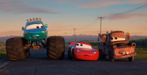 Lightning McQueen (center) and his best friend Mater (far right) from the beloved Cars franchise are joined by a new friend/Monster truck as they travel down a highway at dusk in the animated series Cars on the Road.