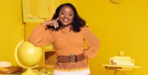 Quinta Brunson, in character as Janine Teagues, wears an orange top and a crocheted skirt. She is resting her right arm on a globe in an entirely yellow classroom.