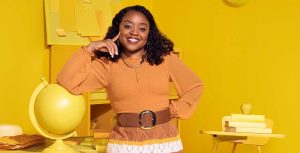 Quinta Brunson, in character as Janine Teagues, wears an orange top and a crocheted skirt. She is resting her right arm on a globe in an entirely yellow classroom.