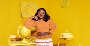 Quinta Brunson, in character as Janine Teagues, wears an orange top and a crocheted skirt. She is resting her right arm on a globe in an entirely yellow classroom.