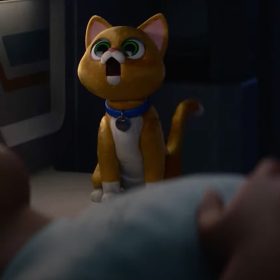 Sox, the feline robot companion from Disney and Pixar’s Lightyear, is seen with his mouth agape, creating “white noise,” as Buzz Lightyear is lying down on a bed in the foreground, sleeping. Sox is a ginger cat with green eyes, and is wearing a blue collar.