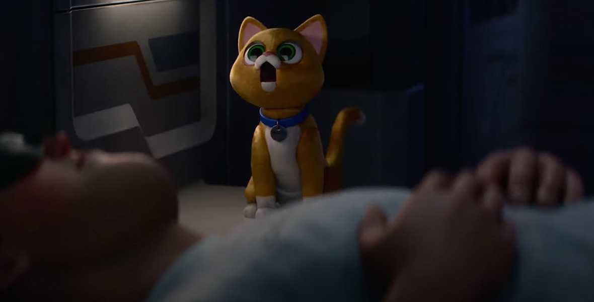 Sox, the feline robot companion from Disney and Pixar’s Lightyear, is seen with his mouth agape, creating “white noise,” as Buzz Lightyear is lying down on a bed in the foreground, sleeping. Sox is a ginger cat with green eyes, and is wearing a blue collar.