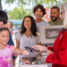 A young girl, left, in a tie-dyed pink T-shirt and rose-gold Minnie Mouse ears is coming through the turnstiles at Disneyland and is getting her MagicBand+ scanned by a Disneyland cast member, right, wearing a long-sleeved red jacket. Behind the girl are other members of her family.