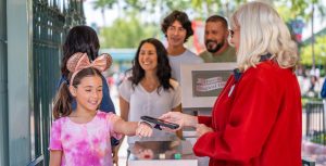 A young girl, left, in a tie-dyed pink T-shirt and rose-gold Minnie Mouse ears is coming through the turnstiles at Disneyland and is getting her MagicBand+ scanned by a Disneyland cast member, right, wearing a long-sleeved red jacket. Behind the girl are other members of her family.