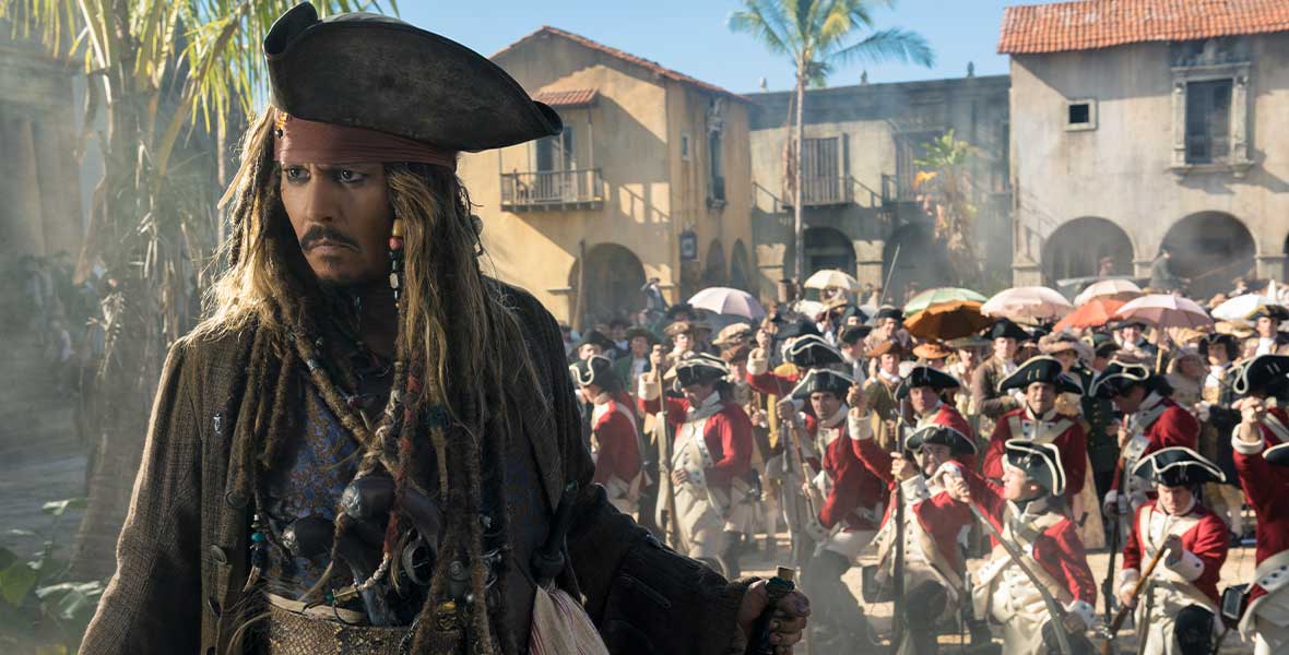 Captain Jack Sparrow wears a brown jacket with two wide belts. His hair is dreaded with miscellaneous beads and ribbons. He is wearing a black pirate hat over a red head scarf. Behind him stand a militia of soldiers in red jackets and white hats.