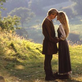 Westley and Princess Buttercup face each other with their foreheads touching and hands held together. They are wearing brown clothing and have blonde hair. They are standing in a pasture overlooking a valley.