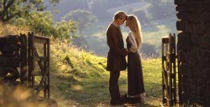 Westley and Princess Buttercup face each other with their foreheads touching and hands held together. They are wearing brown clothing and have blonde hair. They are standing in a pasture overlooking a valley.