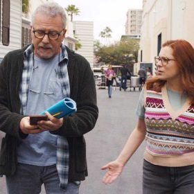 Actors Paul Reiser and Rachel Bloom walk side by side at a film studio backlot. Behind them are large white trailers with bands of blue stripes. Reiser wears jeans and a gray shirt with a plaid shirt open and dark jacket. He wears glasses and holds a script and cell phone in his hands. Bloom wears glasses and holds a script in her left hand. She wears a colorful sweater vest over a gray shirt and black jeans.