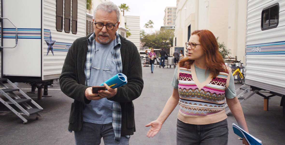 Actors Paul Reiser and Rachel Bloom walk side by side at a film studio backlot. Behind them are large white trailers with bands of blue stripes. Reiser wears jeans and a gray shirt with a plaid shirt open and dark jacket. He wears glasses and holds a script and cell phone in his hands. Bloom wears glasses and holds a script in her left hand. She wears a colorful sweater vest over a gray shirt and black jeans.