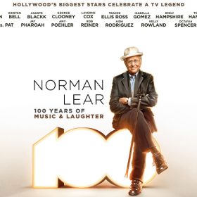In a promotional image for ABC’s Norman Lear: 100 Years of Music and Laughter, Norman Lear is wearing a white hat, a dark sport coat and slacks, and his arms are crossed; his cane is hanging from one arm. He’s sitting on the number “100,” lit up from within. A listing of the stars appearing in the special is seen at the top of the image, and the ABC and Hulu logos are seen at the bottom.