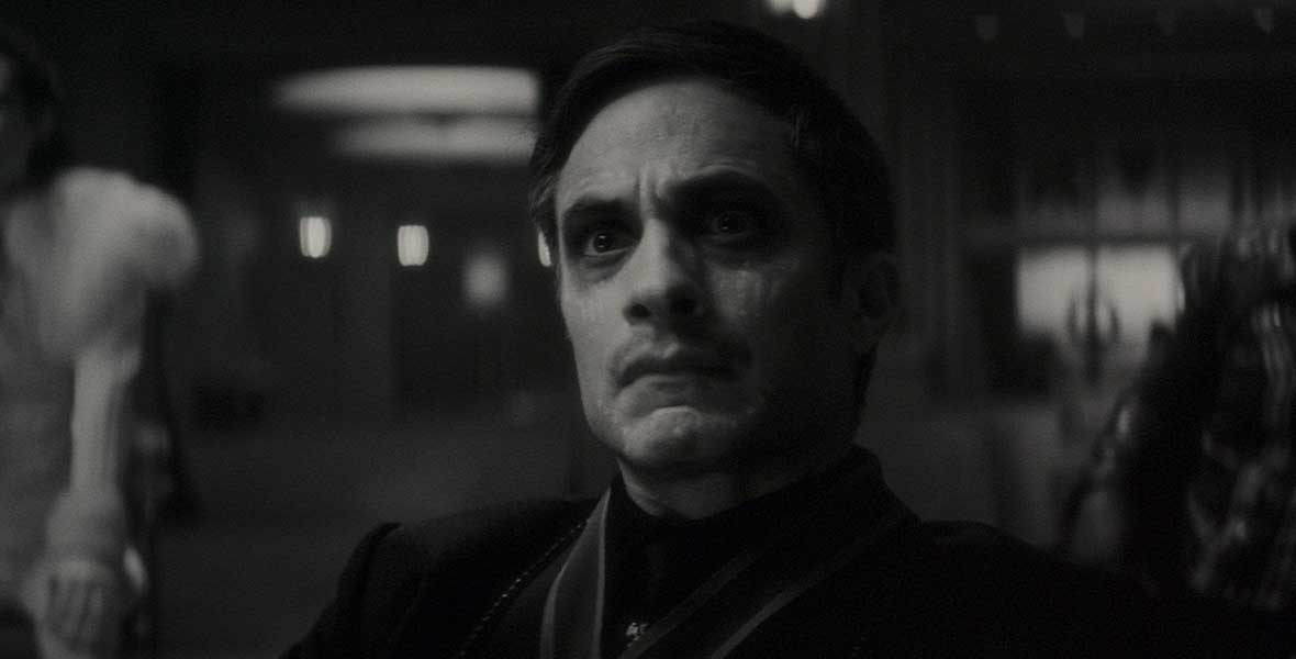 In a black and white scene from Werewolf by Night, Gael García Bernal is seated in a room. He is surrounded by people who are out of focus.