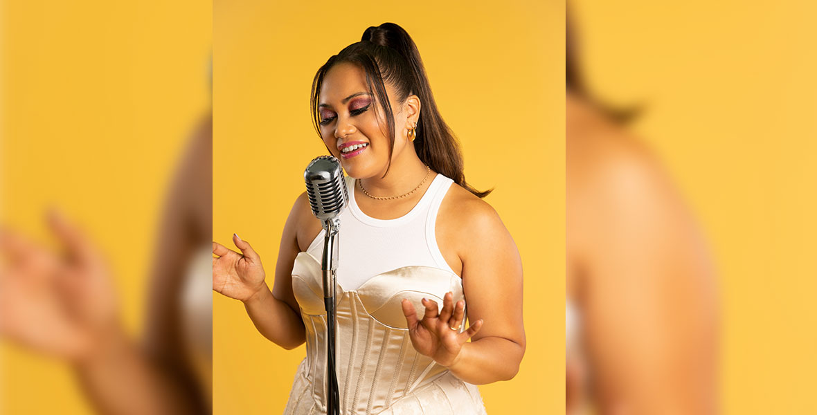 Brittney Mendoza stands against a yellow backdrop and wears a white tank top underneath a corseted cream gown. Her eyes are closed, and she is singing into a microphone with her hands raised.