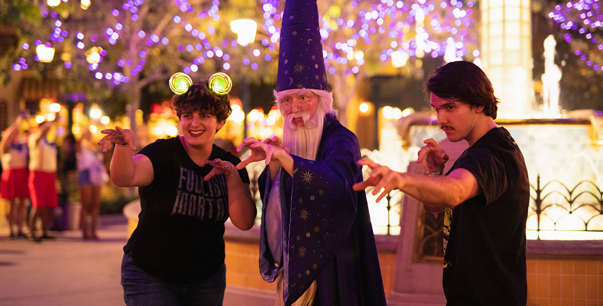 Three guests posing with their best spell-casting poses. The guest on the left is wearing a black Foolish Mortals shirt, light up orange and green Minnie ears and blue jeans. The guest is smiling and has short brown hair. The guest in the middle is dressed up as Merlin from The Sword and the Stone. The guest has a large, blue, pointed wizard hat with silver stars. The guest has silver hair and a long silver beard. The guest is also wearing circular glasses and a long blue wizard robe with silver stars. The guest on the right is wearing a black shirt and black pants. The guest has short brown hair and mustache. Behind the guests to the right is a fountain lit up with yellow lights. In the back are trees lit up with purple lights.