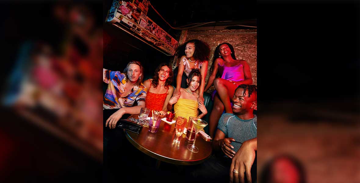 A group photo of Ben Hard, Claude Shwartz, Ebon Gore, Fernando Casablancas, Sophia Wilson, and Taofeek Abijako shows them smiling and wearing solid, brightly colored clothing. They are sitting around a wood table in a corner booth. The lighting is dim, and a brick wall is behind them.