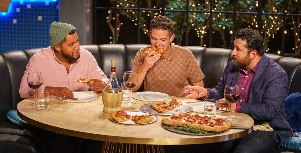 Bryan Ford, Wells Adams, and Daniele Uditi sit around a round wood table placed within a black leather booth. Ford and Uditi talk to each other from across the table. Adams is biting the pizza. On the table are plates and dishes with different slices of pizza.