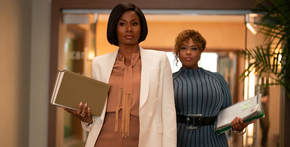 From left to right, actors Emayatzy Corinealdi and Angela Grovey walk through glass doors and down a hallway. Corinealdi wears a white blazer with a tan blouse and tan pants. She holds two large books in her right hand. Grovey wears a blue-gray dress with a large black belt and silver buckle around her waist. The dress has vertical black stripes. She holds files in her right left hand. 