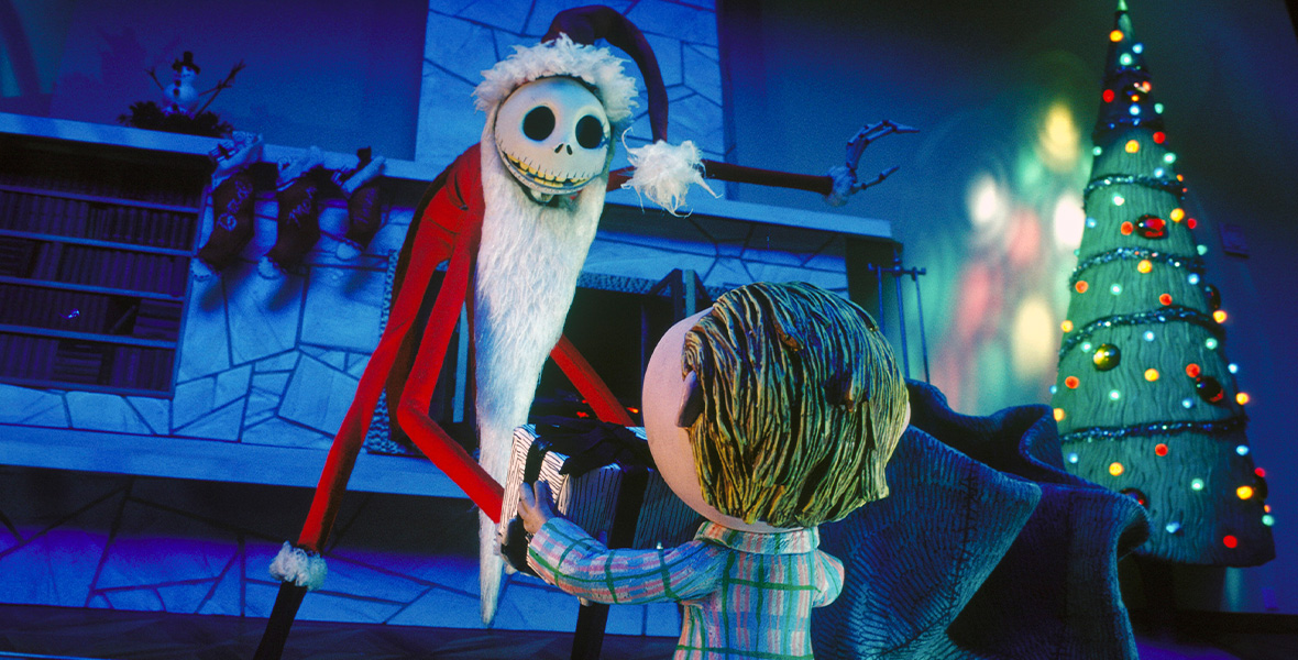 A skeleton-like man wears a red Santa Claus suit and hat, both with white fur piping. Behind him is a stone fireplace with three stockings hanging from the mantle. A young boy faces the skeleton and holds a large present. He wears blue, plaid pajamas. A tall green Christmas tree is in the righthand corner of the room. It is lit with red, yellow, and green lights.