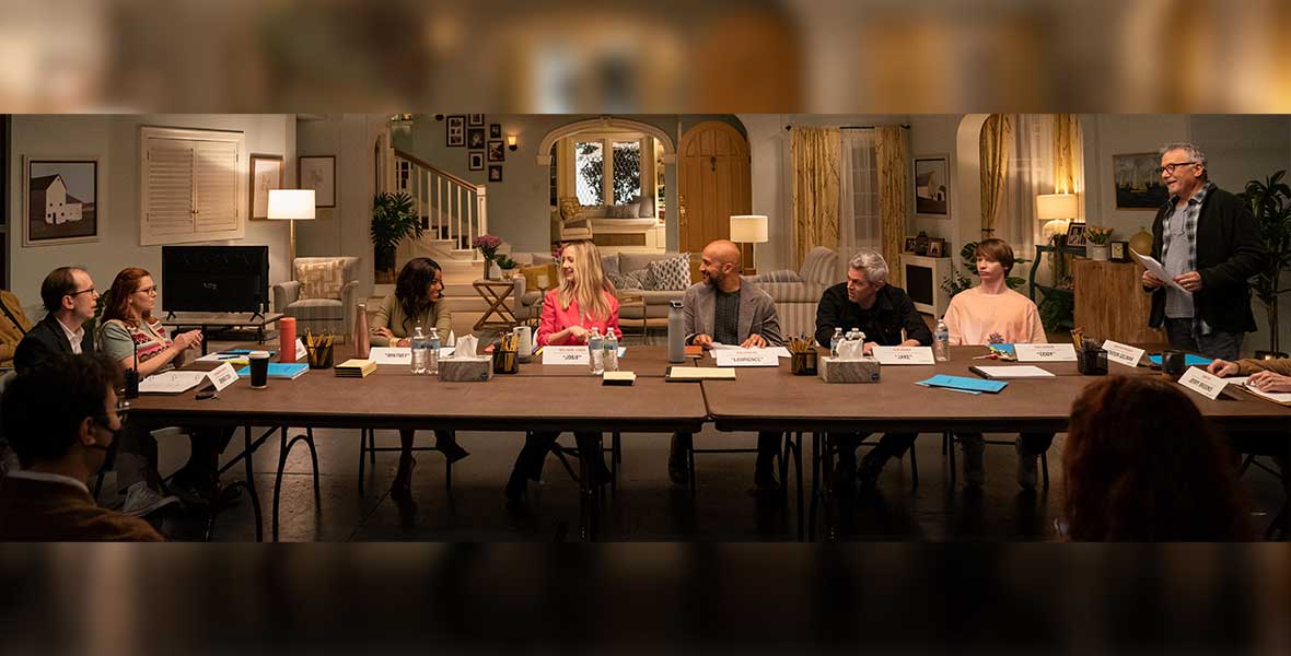 Large tables are placed horizontally in a living room set. Around the table are actors Rachel Bloom, Keegan-Michael Key, Judy Greer, Johnny Knoxville, Callum Worthy, and Paul Reiser. Each have name plates on the table in front of them reading their character names along with scripts. Also on the table are boxes of tissues, bottles of water, and black containers holding dozens of pencils and pens.