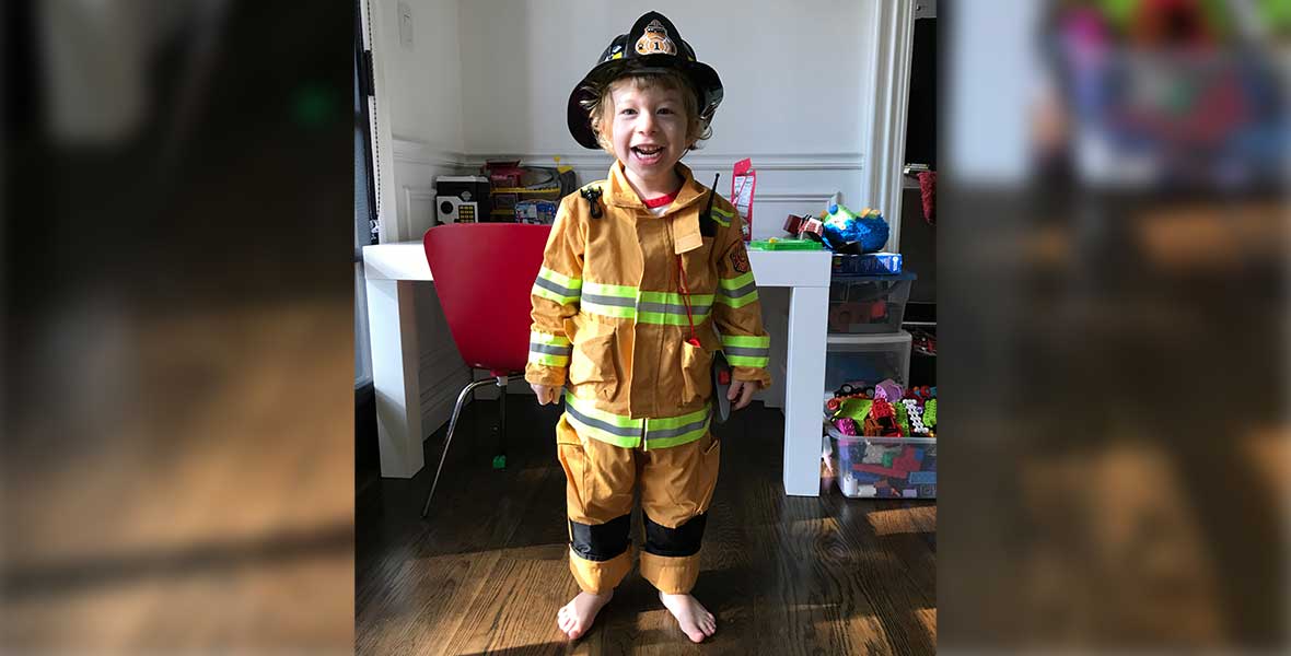 A young boy smiles and stands in front of a white desk. He wears a yellow firefighter suit and black firefighter helmet.