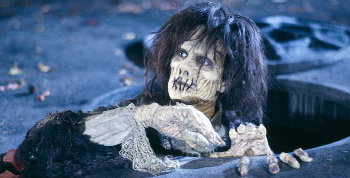 In a still from Hocus Pocus, zombie Billy Butcherson, played by Doug Jones, crawls out of a manhole. His lips are sewn shut, his long black hair is gnarled, and his 17th century clothes are tattered.