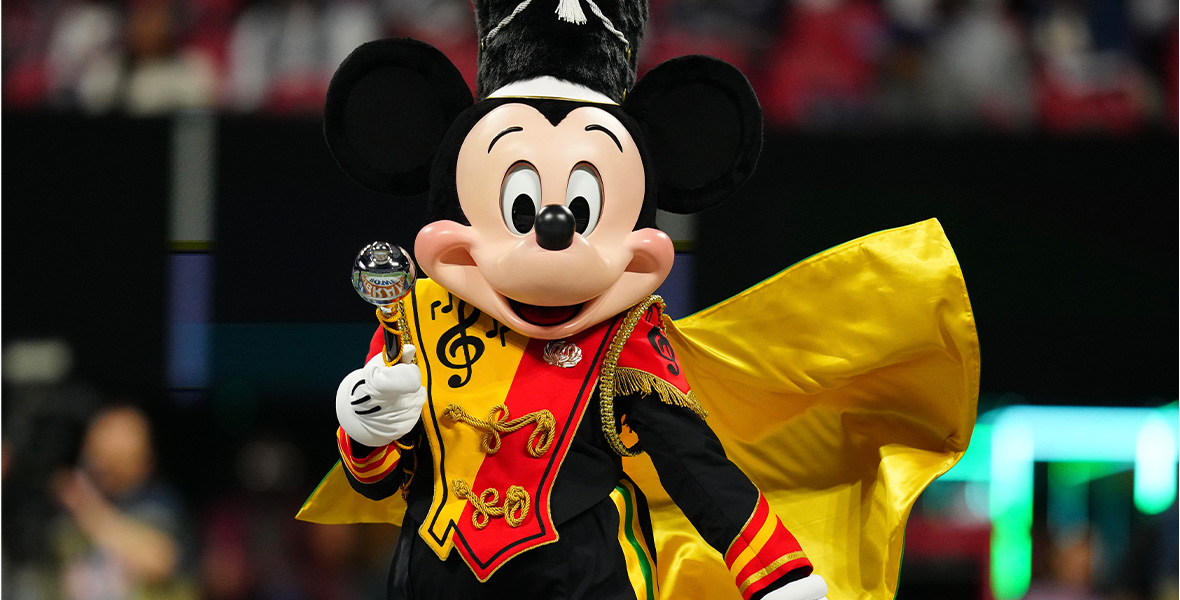 An image of Mickey Mouse in a drum major uniform—the jacket is yellow and red, with a treble clef and music notes and gold knots across the front, and a gold cape flying out from his shoulders. He’s wearing a fuzzy black drum major hat, and is carrying a drum major baton.