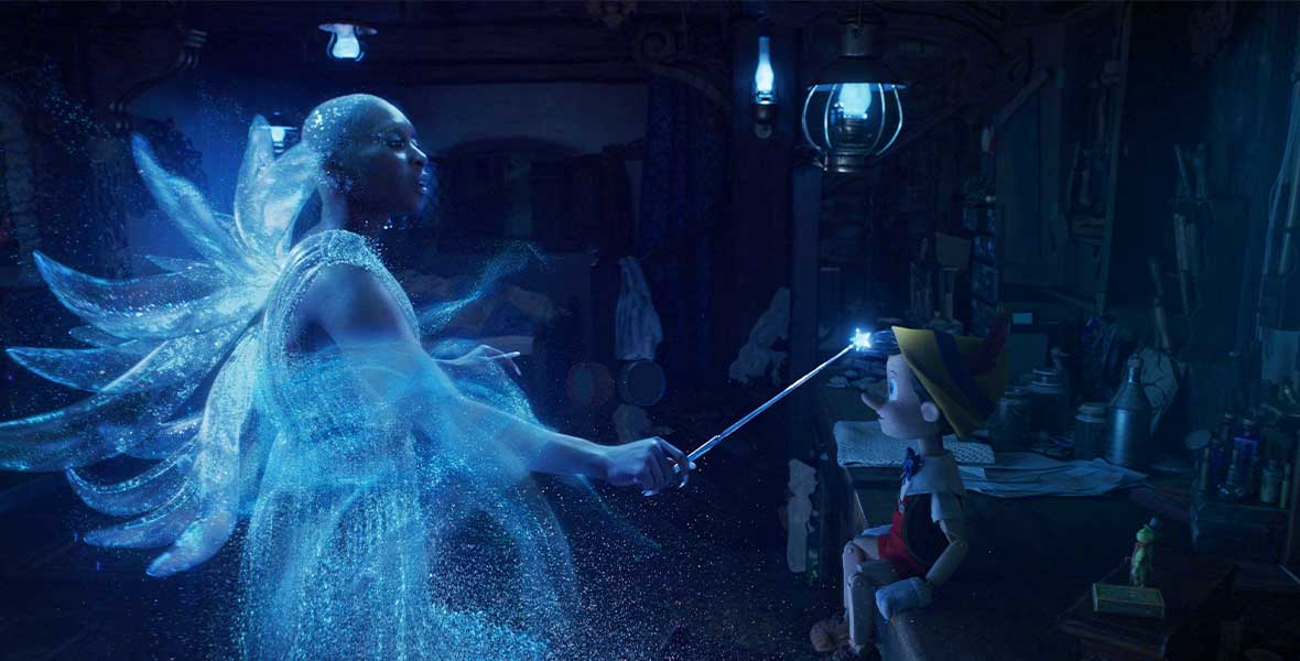 The Blue Fairy, played by Cynthia Erivo, shimmers in blue light as she waves her magic wand in front of the wooden puppet Pinocchio, voiced by Benjamin Evan Ainsworth. Pinocchio is seated in Geppetto’s dimly lit woodshop, and Jiminy Cricket is standing behind him, watching the magical moment unfold.