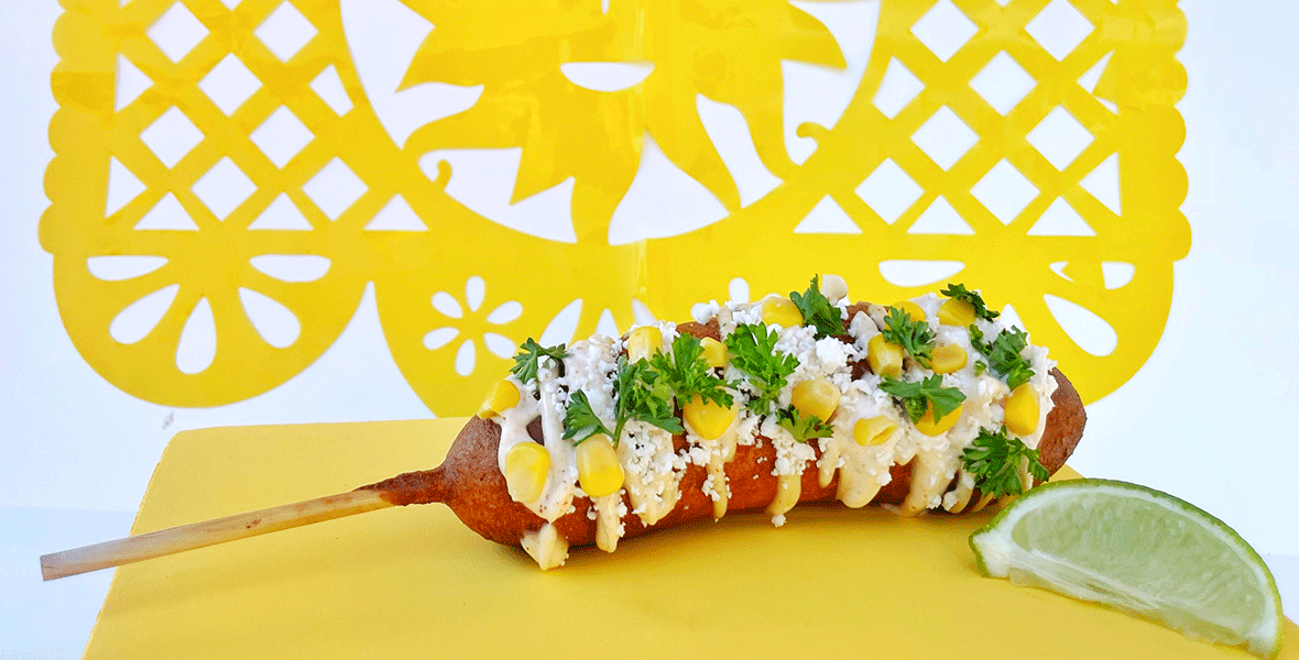 Spicy Hot Link Sausage, dipped in corn meal batter, fried, topped with garlic mayo, corn kernels, cotija cheese, and cilantro laying in front of a bright yellow background.