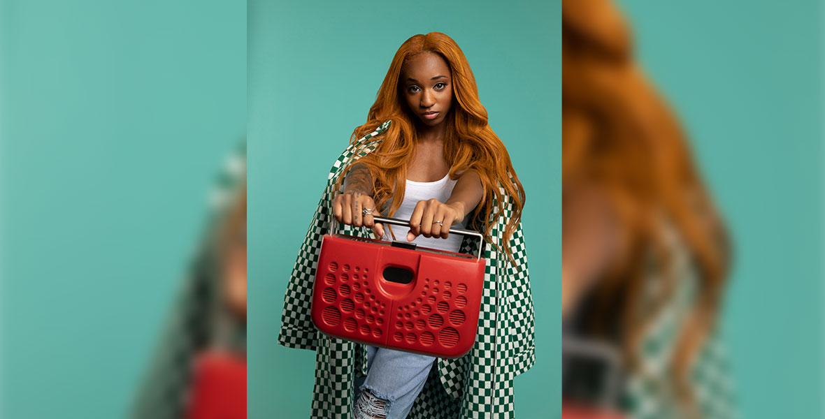 Gabby Samone stands against a green backdrop and wears a checkered jacket, distressed denim pants, and a white tank top. Her arms are outstretched towards the camera, and she is holding a red boombox. 