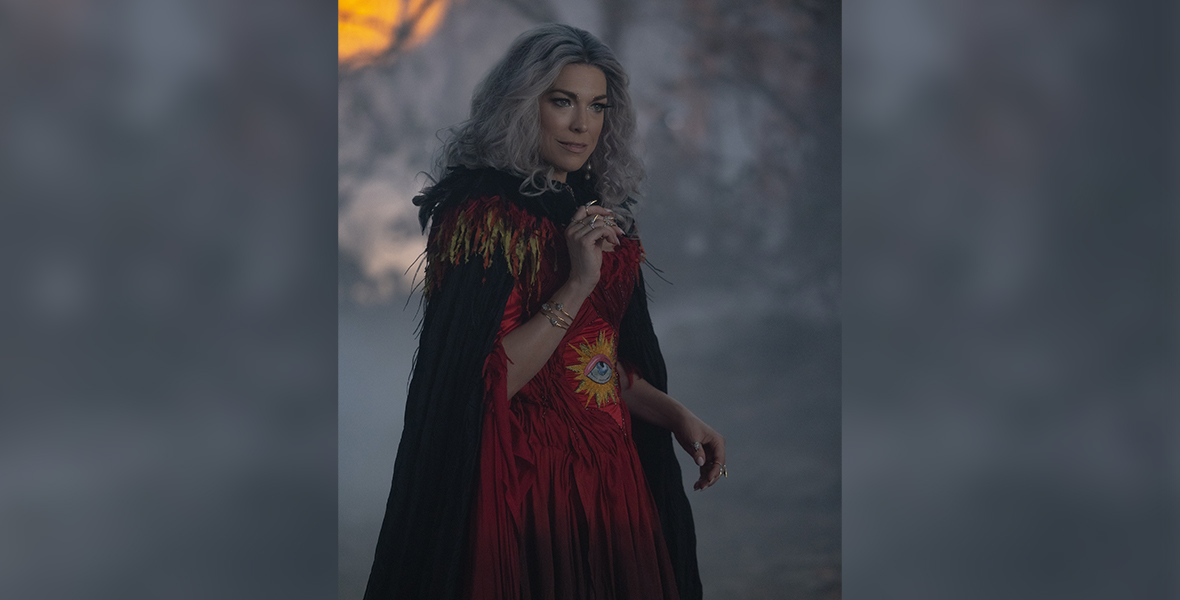 Hannah Waddingham, in character as The Witch Mother, stands alone in the Forbidden Woods of Salem. She wears a crimson red dress, which has a pleated skirt, as well as a print of an eye inside a sunburst on her midriff. Her black cape is feathered and includes red and yellow feathers around the color. She has her right arm raised just below her chain, exposing her gold jewelry. She has a curious expression on her face.