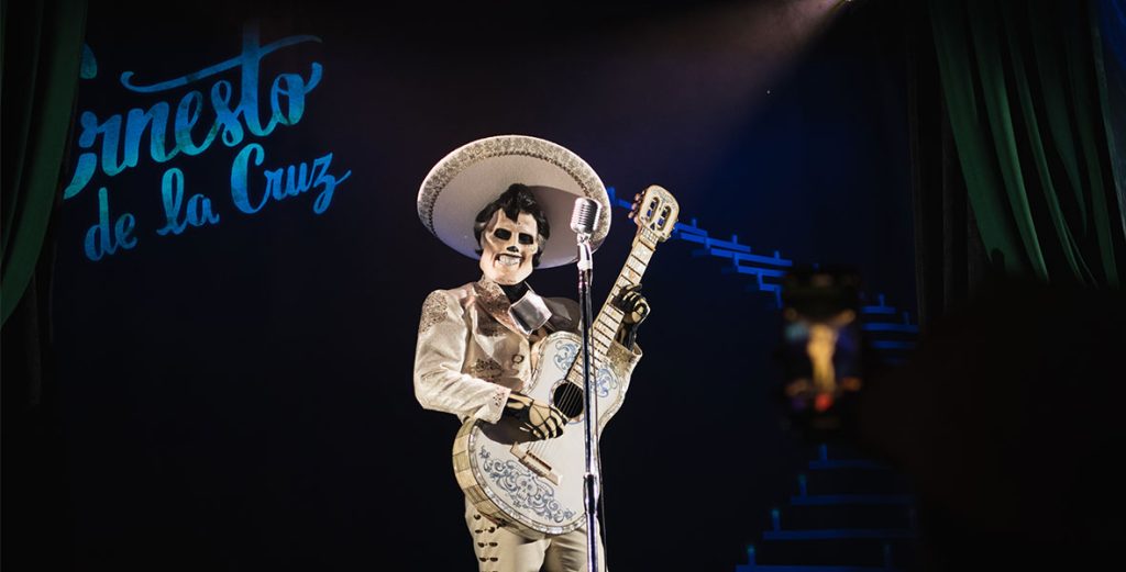 Ernesto De La Cruz smiling with his white guitar and stand-up microphone. Ernesto has black, styled hair with a white skeleton face and hands. Ernesto is wearing a white mariachi suit with a white and gold sombrero, white and gold detailed jacket and pants. To the back left of Ernesto, is Ernesto De La Cruz written and projected in blue.