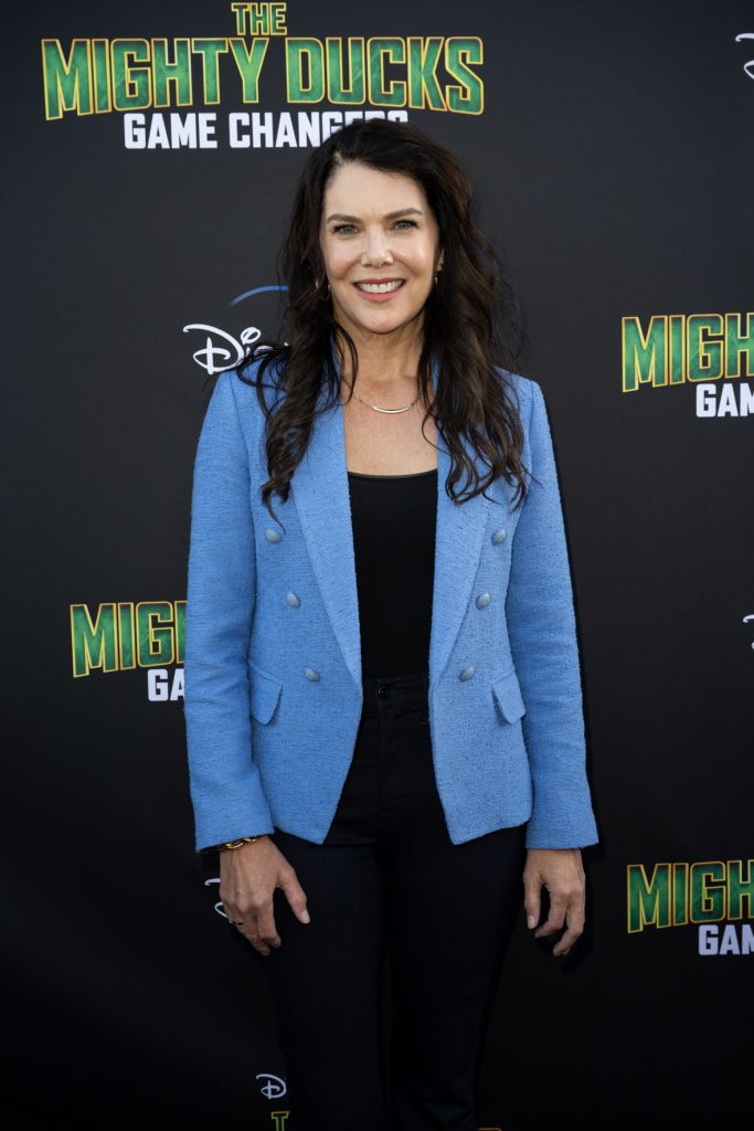 Actor Lauren Graham stands on the purple carpet at the premiere event for the Season 2 The Mighty Ducks: Game Changers. She wears a black top and pants with a light blue blazer jacket.