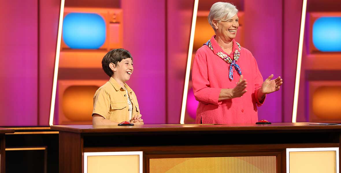 Contestants Paxton and Cindy smile and stand behind a large wood desk. Paxton is a young boy and wears a yellow, short-sleeve shirt. Cindy is an older woman and wears a pink long-sleeve blouse and colorful scarf tied around her neck. A digital screen reads, “Paxton and Cindy” on the desk.