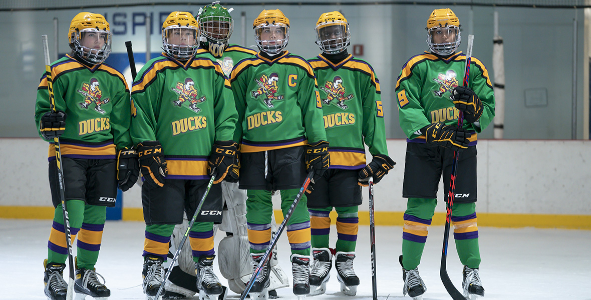 From left to right, actors Taegan Burns, Maxwell Simkins, Luke Islam, Brady Noon, DJ Watts, and Swayam Bhatia stand inside on a hockey rink. Each wear green hockey jerseys with yellow helmets, black shorts, and black skates. All jerseys have a skating duck logo and text reading “DUCKS” across the front of the jersey.