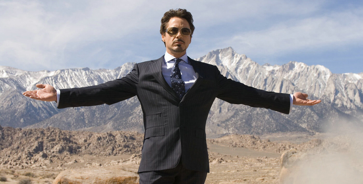 Disney Legend Robert Downey Jr. stands with his arms extend outward. He is surrounded with desert-like terrain with a large mountain range seen in the distance. He wears a dark blue suit with a white pinstripes, blue patterned tie, and white shirt. He also wears large sunglasses.