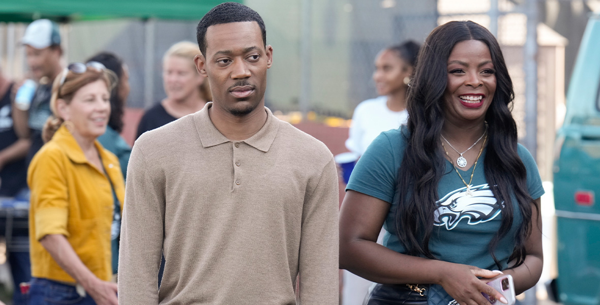 Tyler James Williams, in character as Gregory Eddie, stands with both arms at his side outside of Abbott Elementary. He is wearing a long-sleeved khaki shirt, which is tucked into his jeans. To his left is Janelle James, in character as Ava Coleman, who is holding her iPhone in both hands. She is wearing a Philadelphia Eagles V-beck and a black leather skirt.