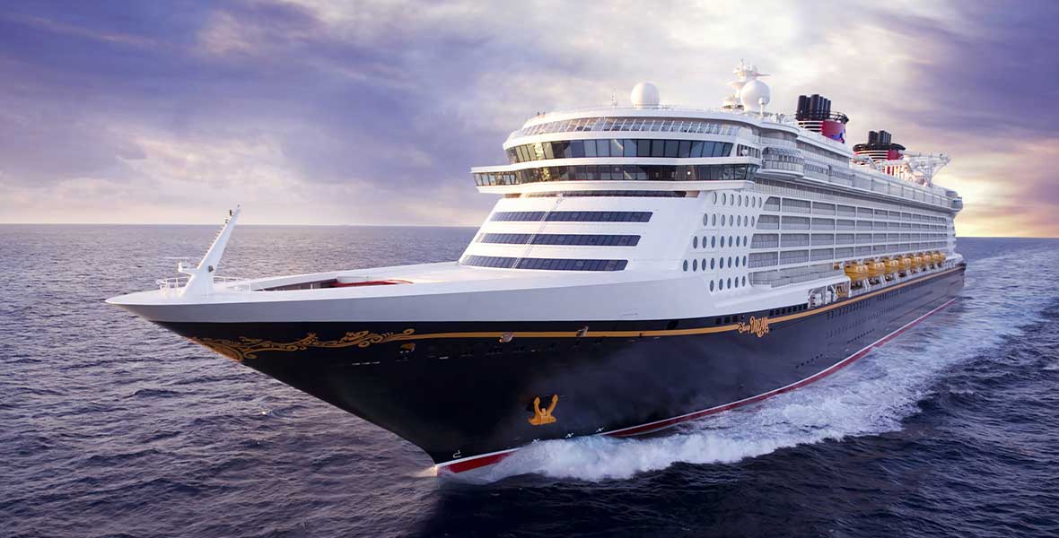 An image of the Disney Dream cruise ship sailing in open water. It’s set against a blue-sky background, with some whispy clouds. The ship is churning up some water toward the bow, as it’s moving through the ocean.