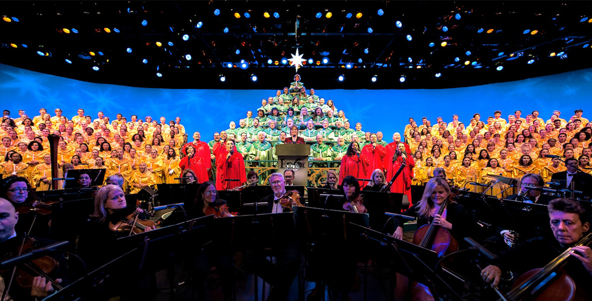 In a promotional image for EPCOT’s Candlelight Processional, a large choir—decked out in yellow, green, and red robes—is seen on stage, behind an orchestra. The choir members in green robes are on risers so as to depict a “human” Christmas tree. A woman is at a lectern, telling the Christmas story. The background on stage is blue with faint white snowflakes, and at the top of the image you can see many stage lights hanging from the rafters.