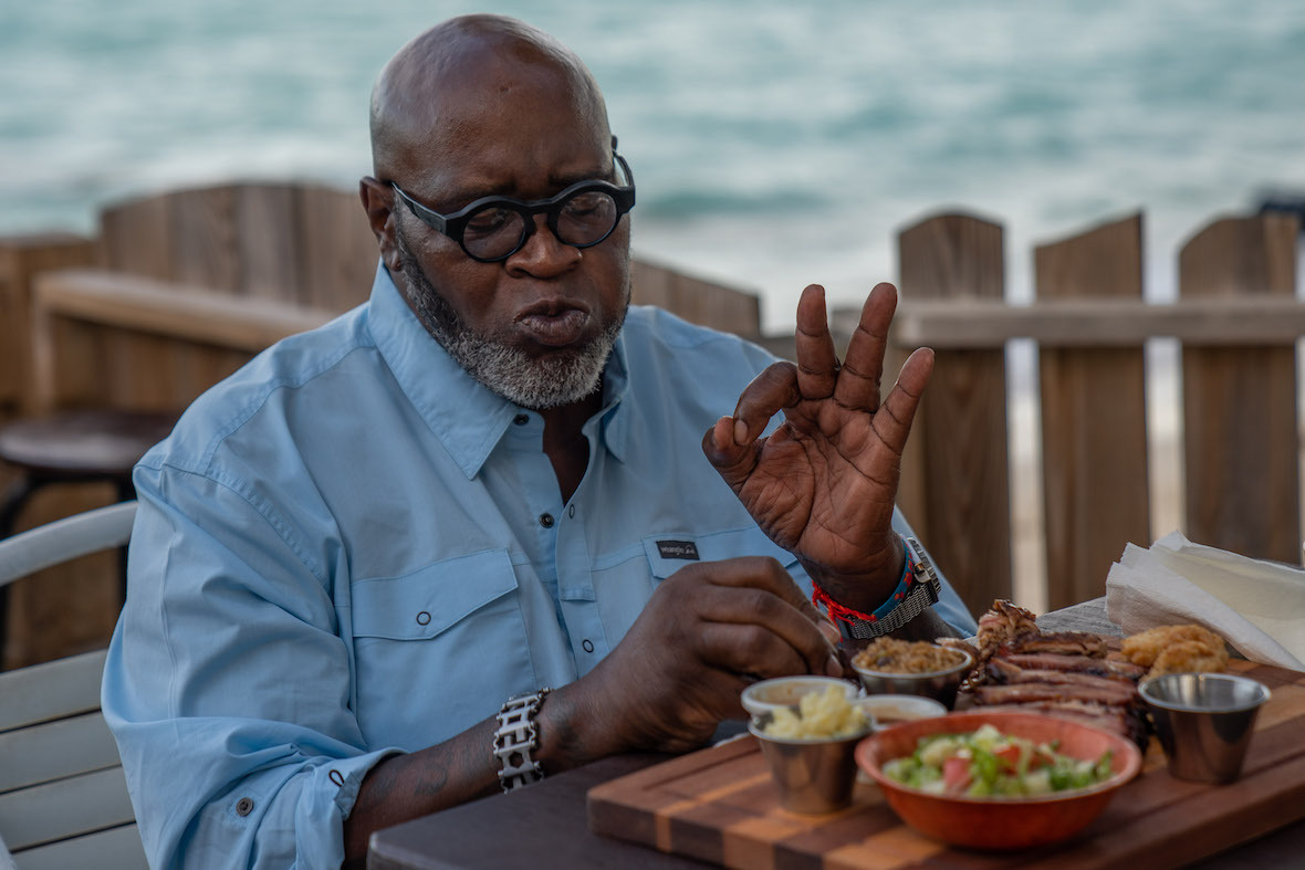 Big Moe Cason enjoys a meal by the water. He is wearing circular glasses, a blue Wrangler shirt, and watch. While pursing his lips, he makes the A-OK symbol with his right hand.